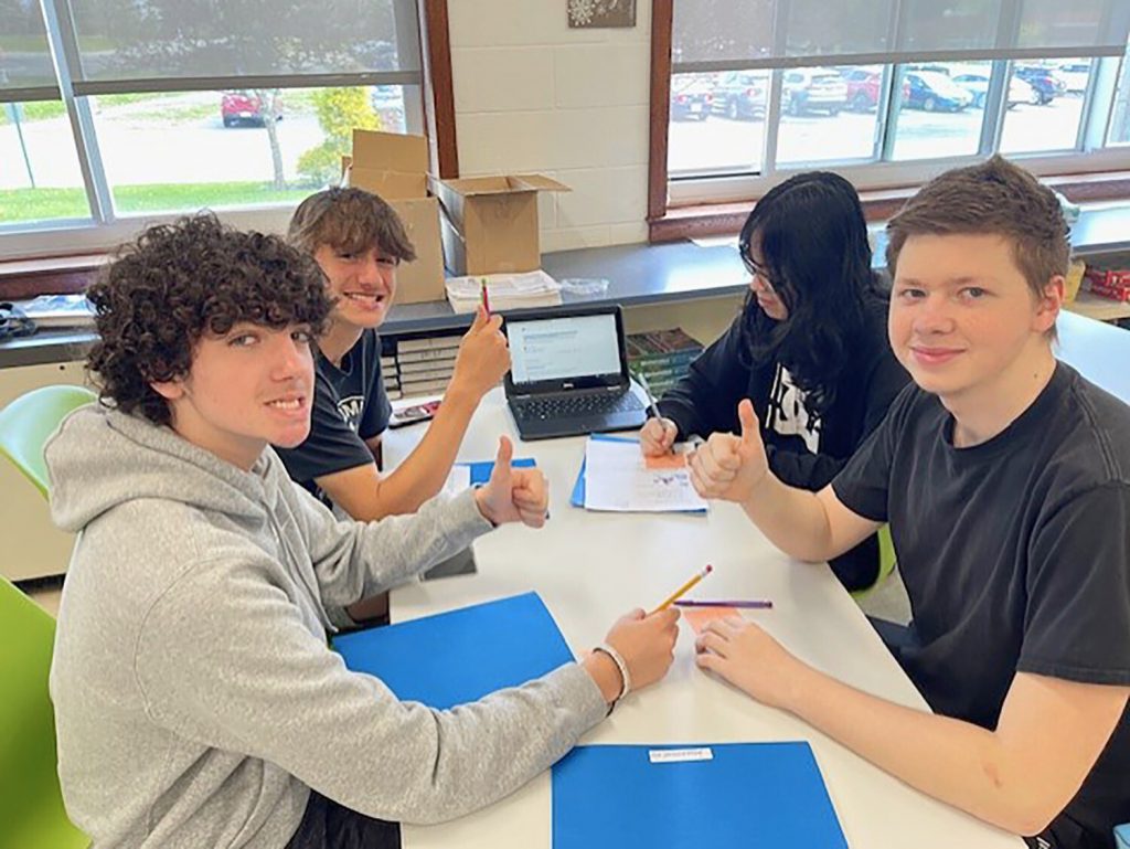 Four students sitting together at a table. Three students are looking at the camera and smiling and the fourth student is writing on a piece of paper and looking down at the table.