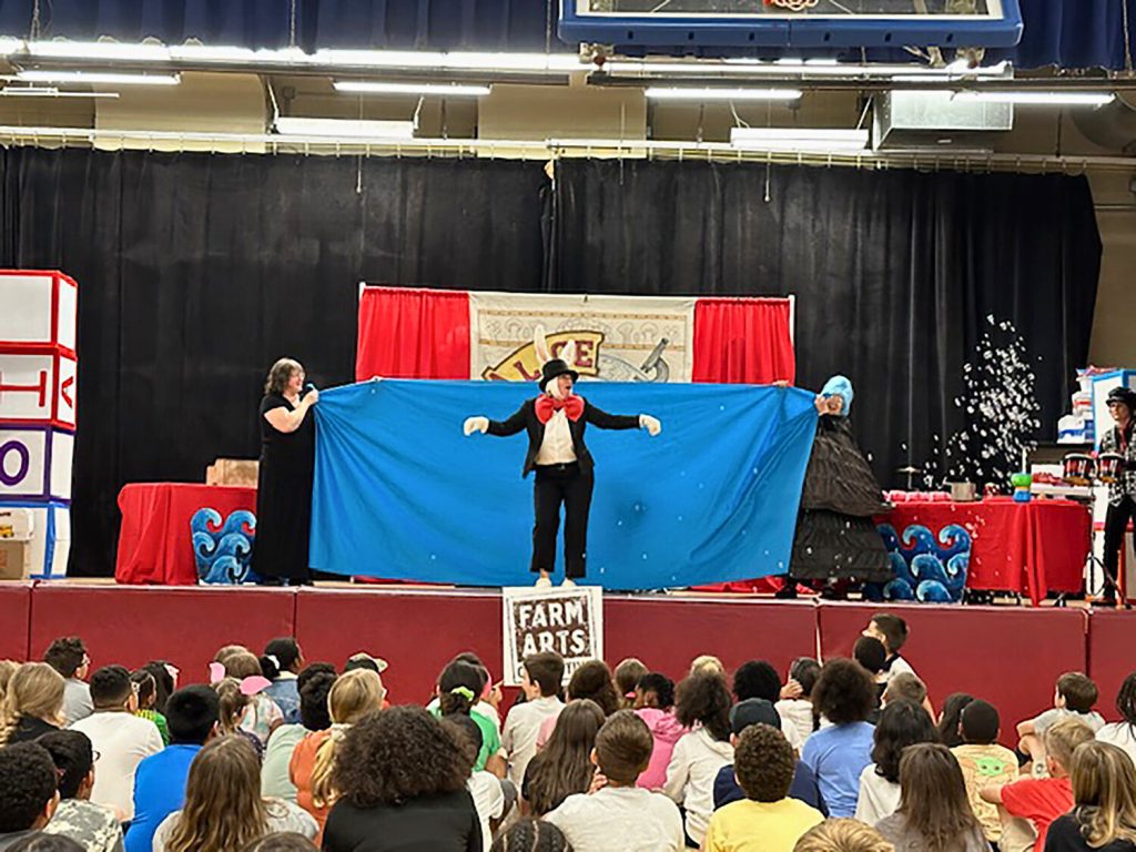 Three performers on stage. The person in the middle is dressed as a rabbit. Two people are standing on each side of the middle performer and they're holding a blue sheet.