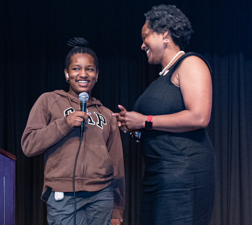 a student is holding a microphone and speaking to an audience as an adult woman looks on and smiles 