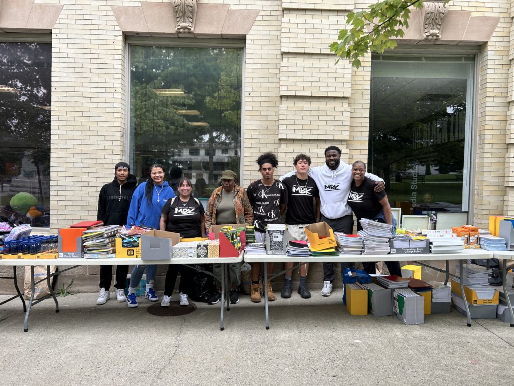 a group of MBK members are standing in front of a table filled with school supplies. 