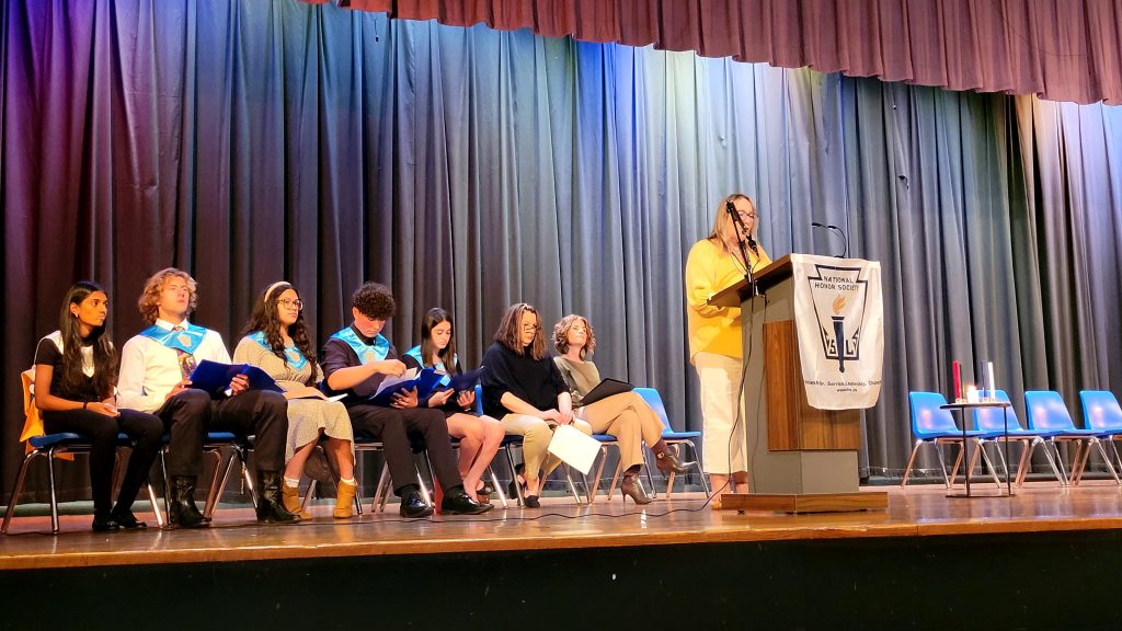 National Honor Society inductees are seated on stage. A speaker is at the podium addressing the audience. 