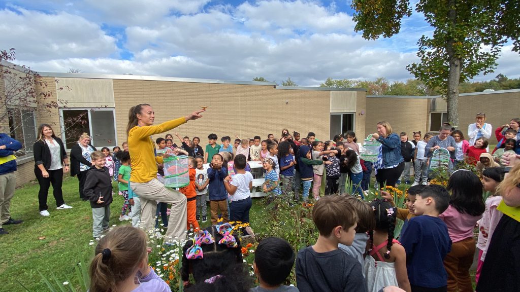 a teachers is addressing a large group of students outdoors. The students are gathered around her. 