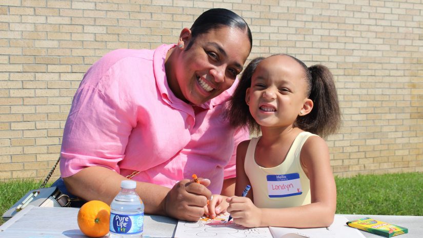 a mom is sitting at a table with her daughter. They're smiling for the camera with a coloring book in front of them.