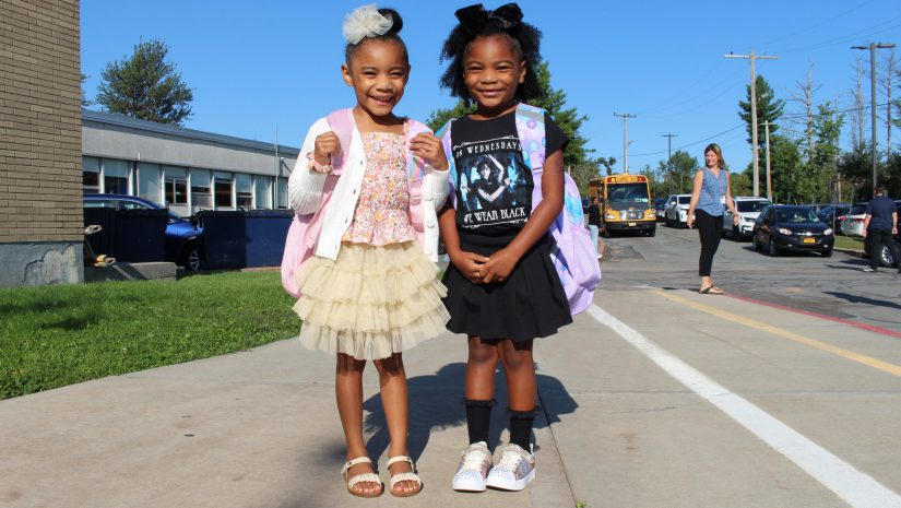 two young girls with backpacks are posing in front of the school