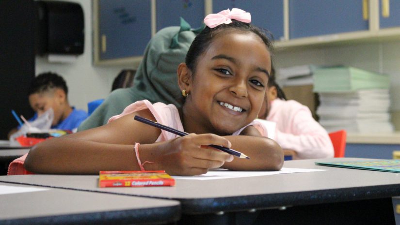 a young girl seated at a desk with a pencil in her hand is looking up at the camera and smiling