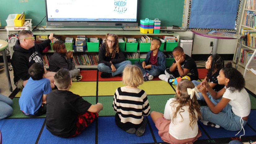 a group of students and their teacher are in the middle of a lesson. They are seated in a circle on a rug