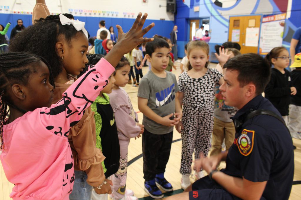 a firefighter is speaking to students. One student has her hand raised, about to ask a question.