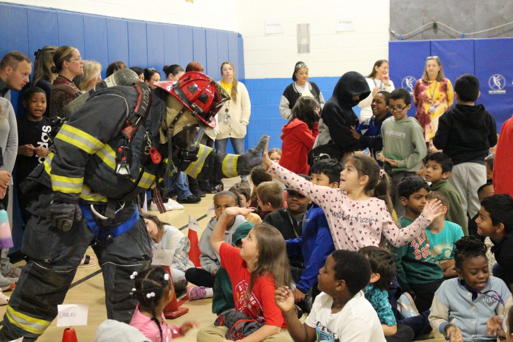 A firefighter in full gear is surrounded by a group of students. One is giving him a high five