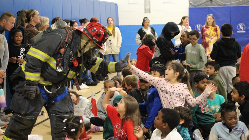 A firefighter in full gear is surrounded by a group of students. One is giving him a high five