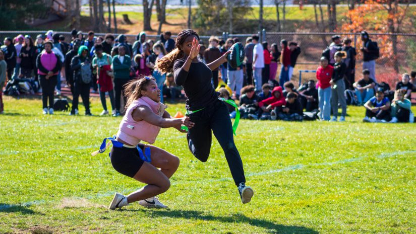 two female students are engaged in a powder puff football game. Once is jumping in the air about to catch a football and the other stands ready to tackle her.