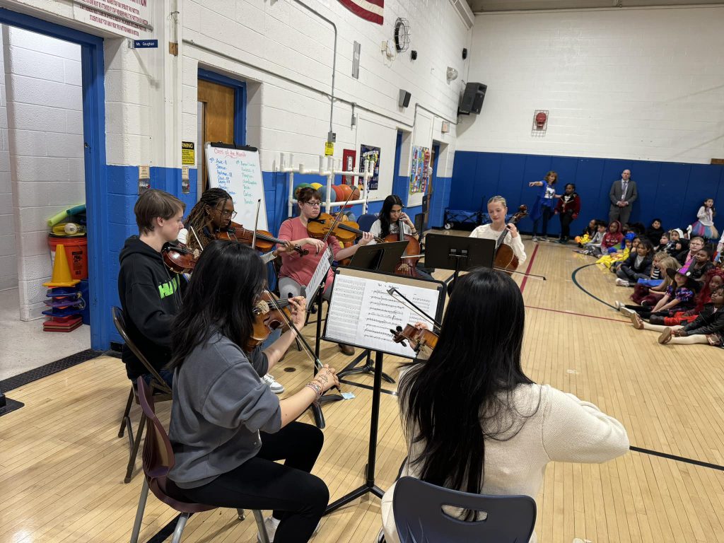 a string ensemble is performing in front of a large group of elementary school students