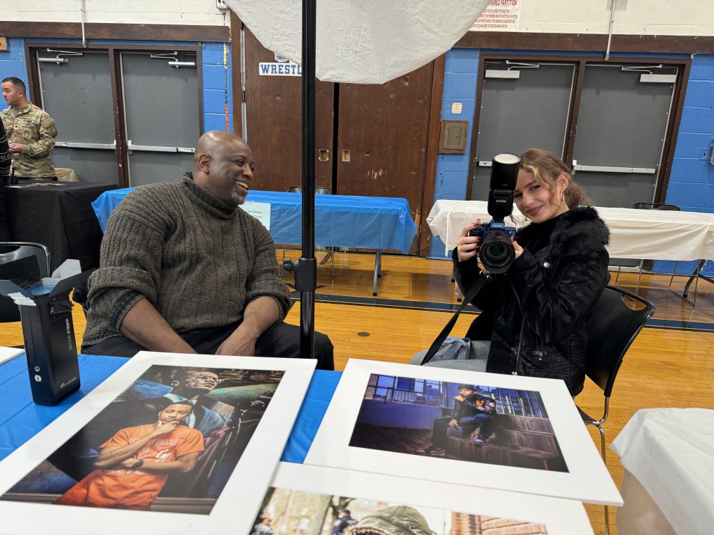 a students is seated with a photographer. She is holding a camera and playfully pointing it at the person taking her photo