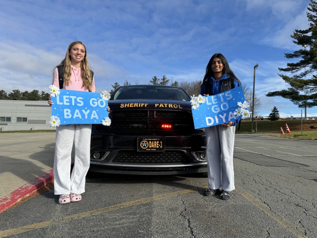 Baya Titus and Diya Patel are posing in front of a police car. They are holding signs that say "let's go Baya and let's go Diya"