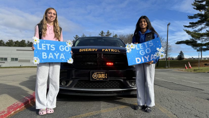 Baya Titus and Diya Patel are posing in front of a police car. They are holding signs that say "let's go Baya and let's go Diya"