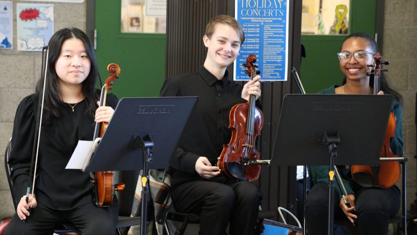 three students seated and holding violins are smiling at the camera