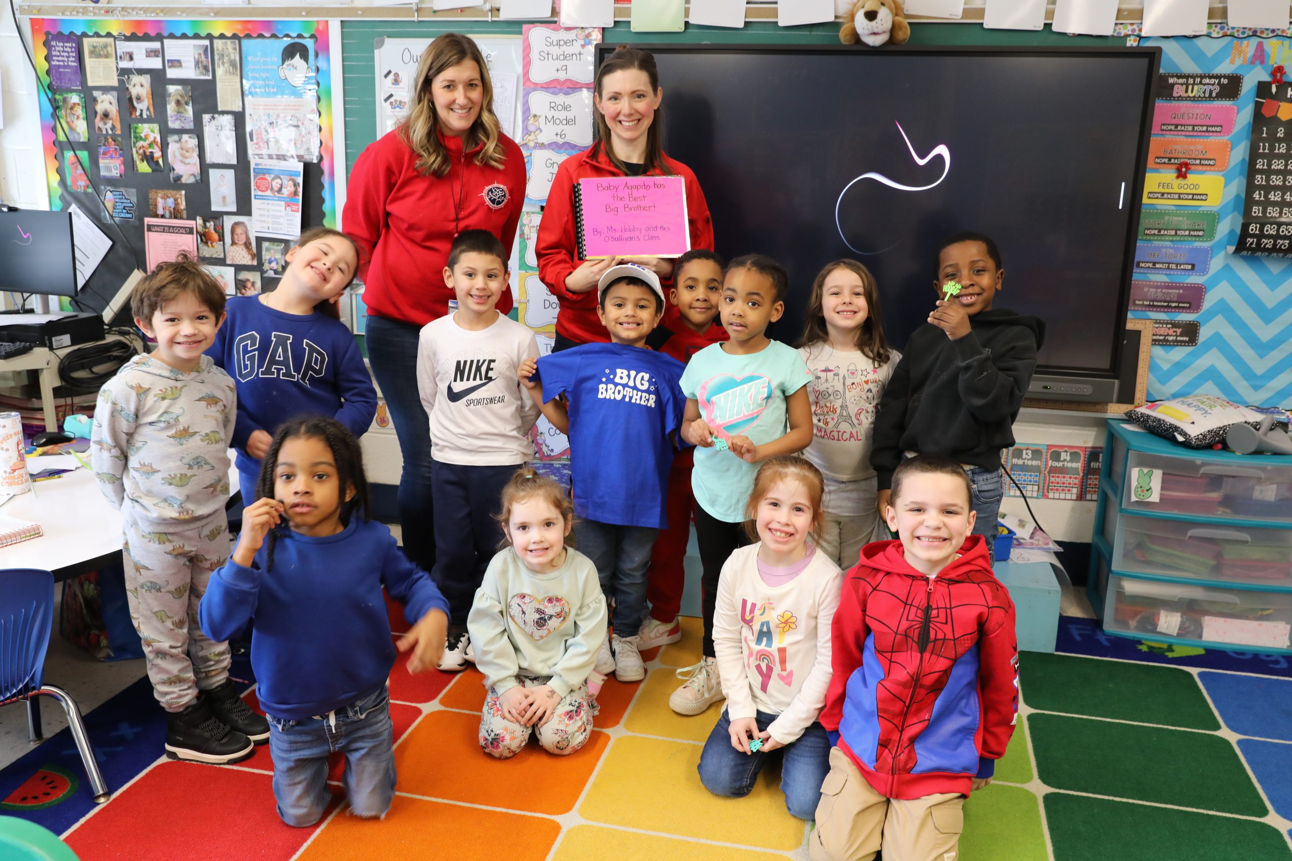 a class full of kindergartners are posing for the camera. The teachers are in the background and one is holding up the book that the class collectively created for their classmates.