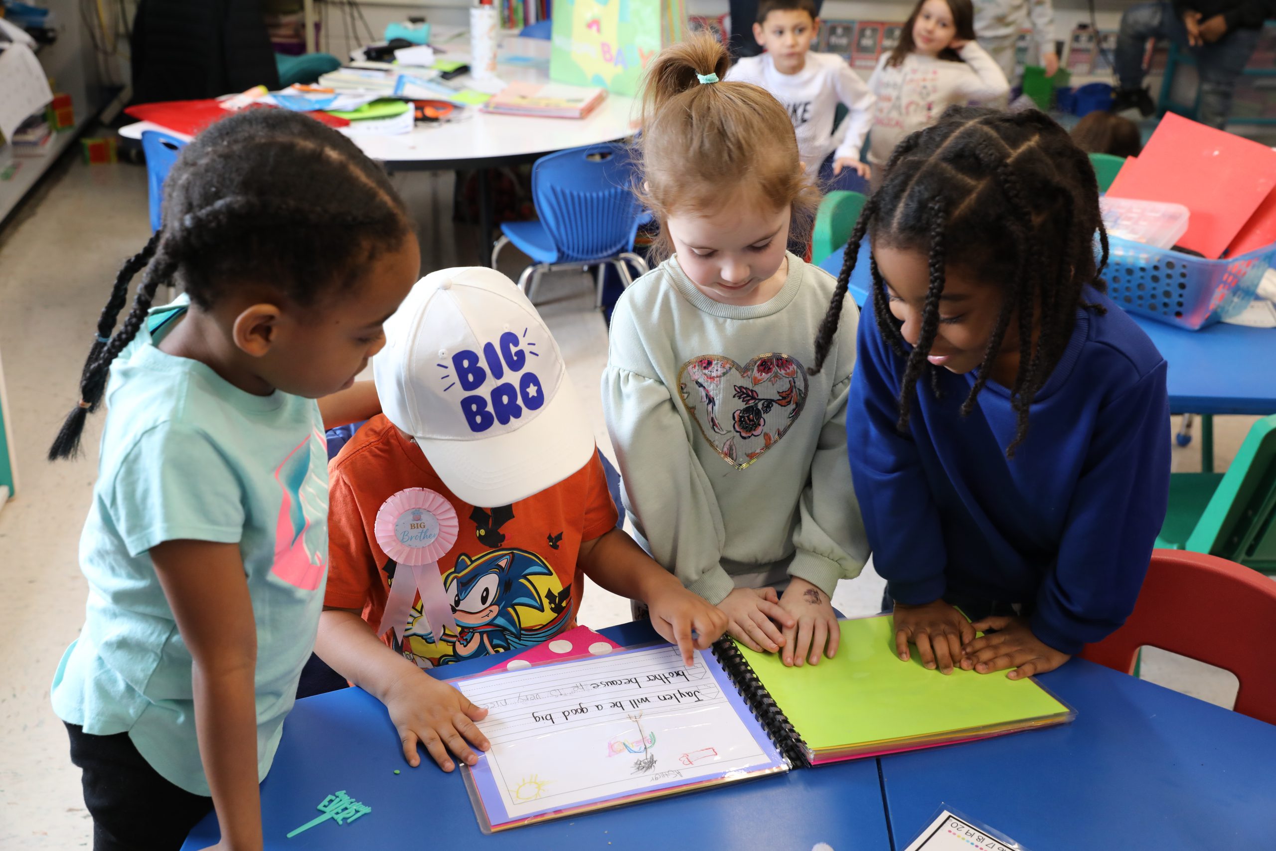 Jaylen, wearing a hat that says big bro is surrounded by a few classmates. They are all looking down and reading a book. 