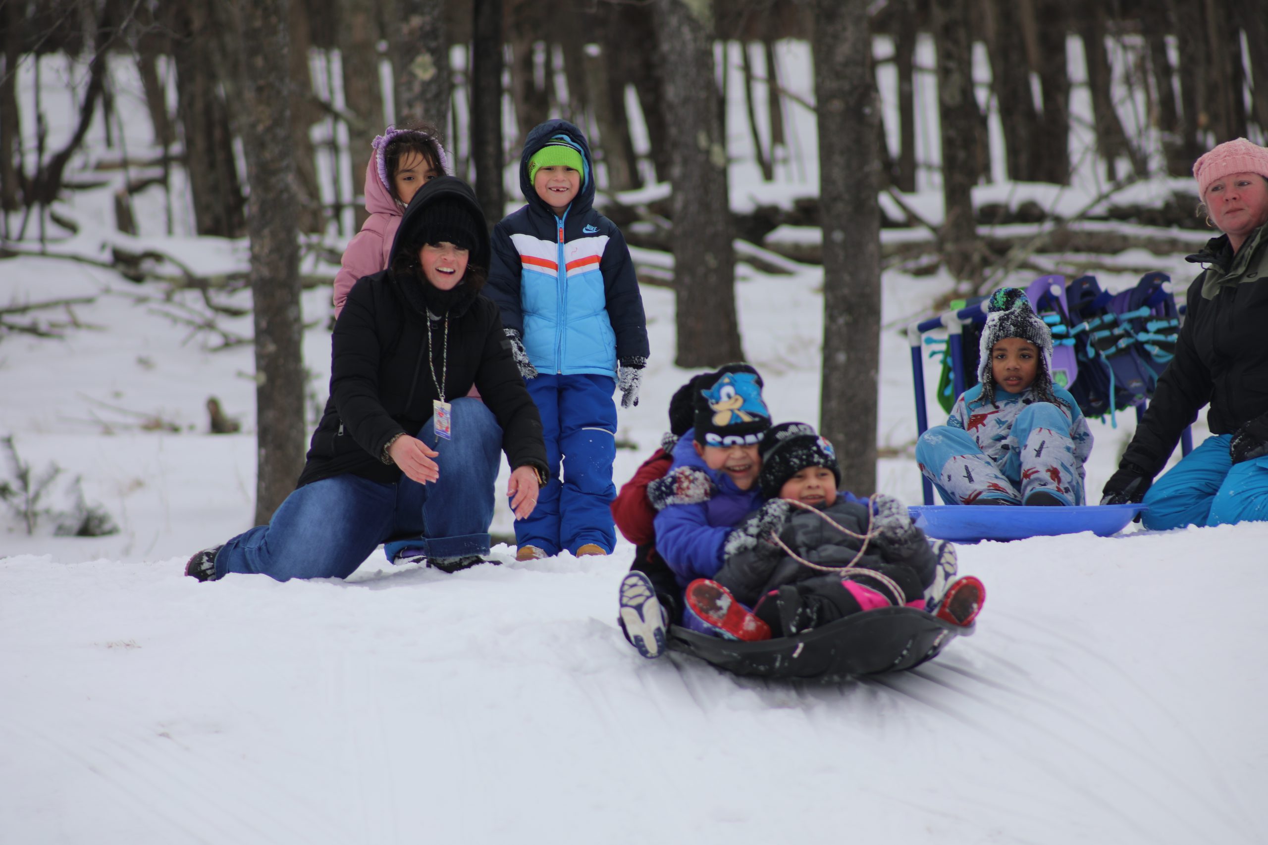 a teacher is pushing a group of three students on a sled down the hill. The students are laughing and smiling. 