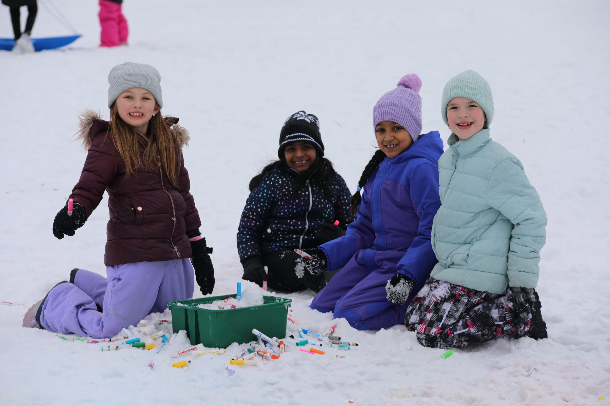 four students are seated in the snow in a circle, using colored water to "color" the snow. 