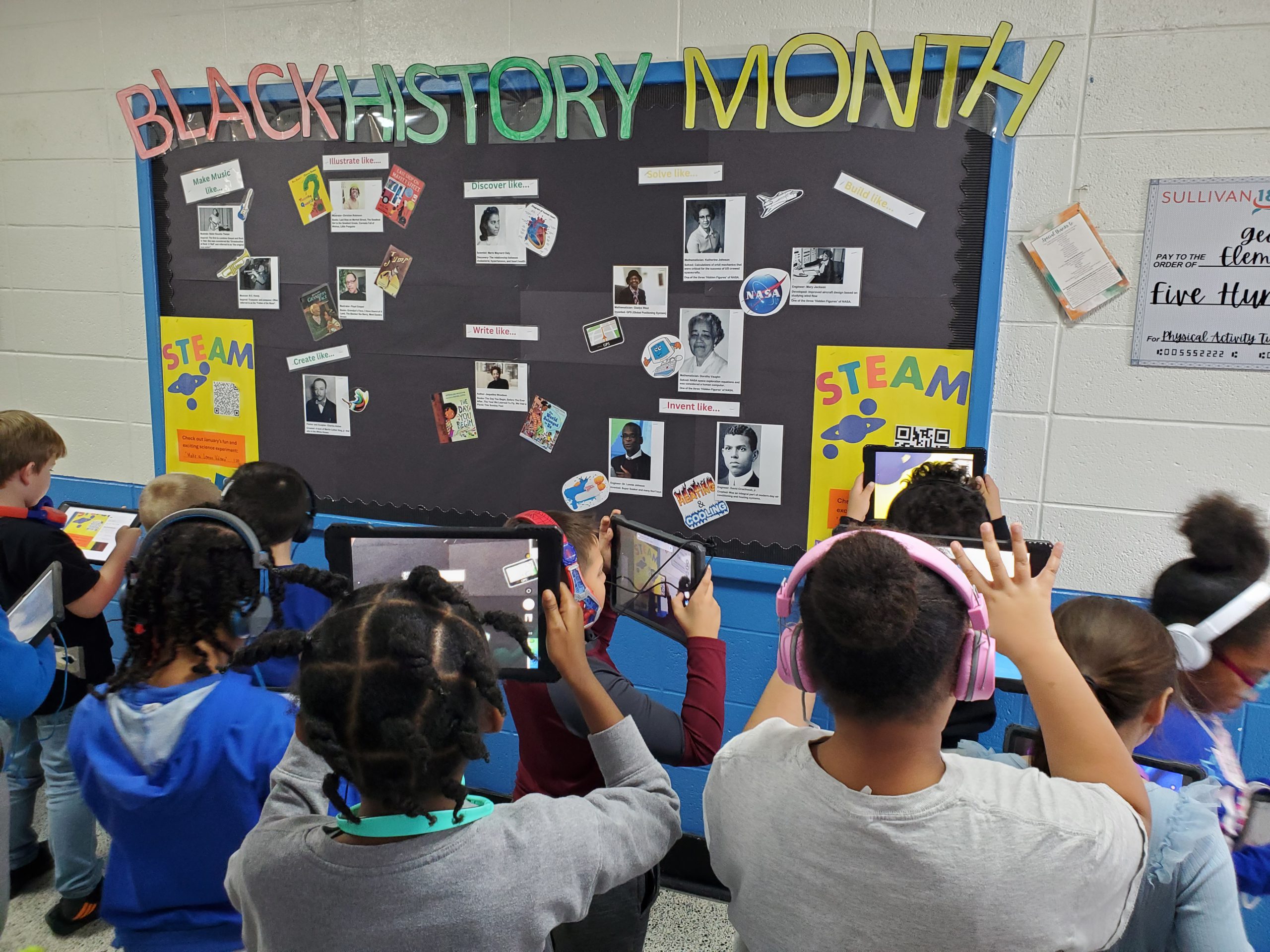 a group of students are holding up iPads towards a bulletin board that says "Black History Month" They are capturing the QR code with their device so that they can learn more about the innovators pictured on the board. 