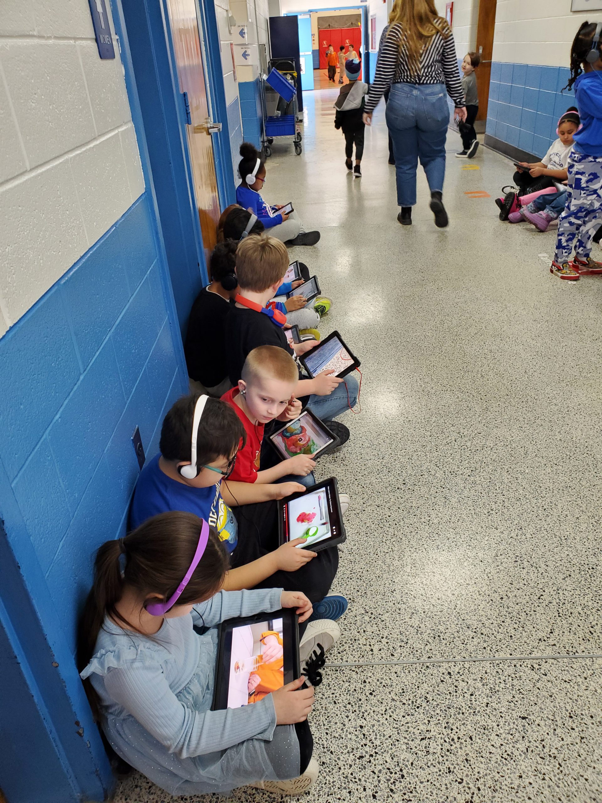 a group of students are seated in a line down the hallway. They are wearing headphones and looking down at their devices, watching videos of the innovators on the bulletin board. 