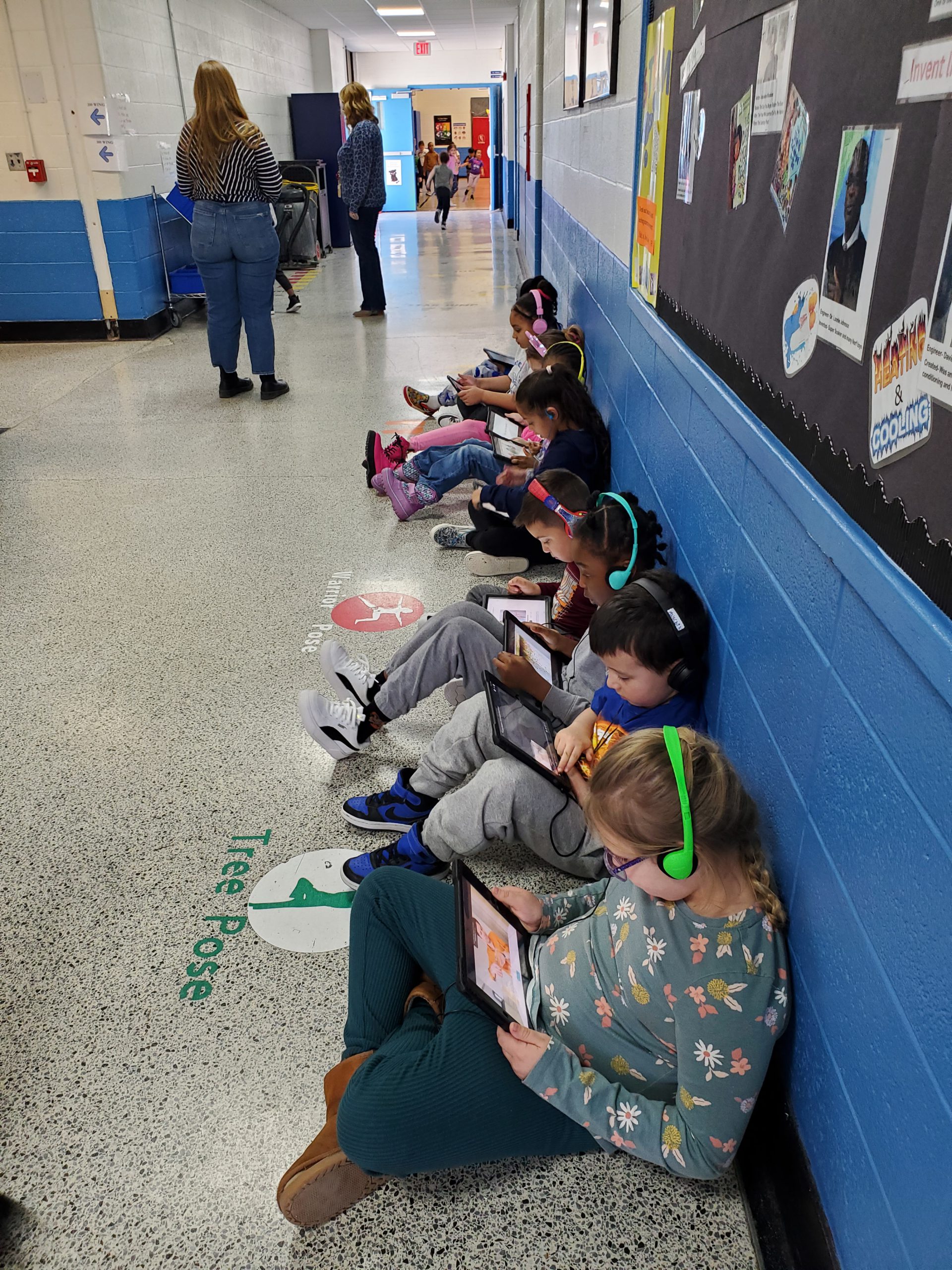 a group of students are seated in a line down the hallway. They are wearing headphones and looking down at their devices, watching videos of the innovators on the bulletin board. 