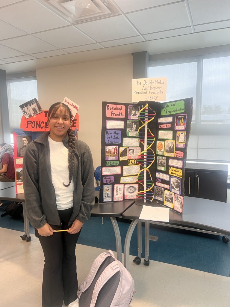 a student poses next to a display of her research project during the local National History Day competition. 