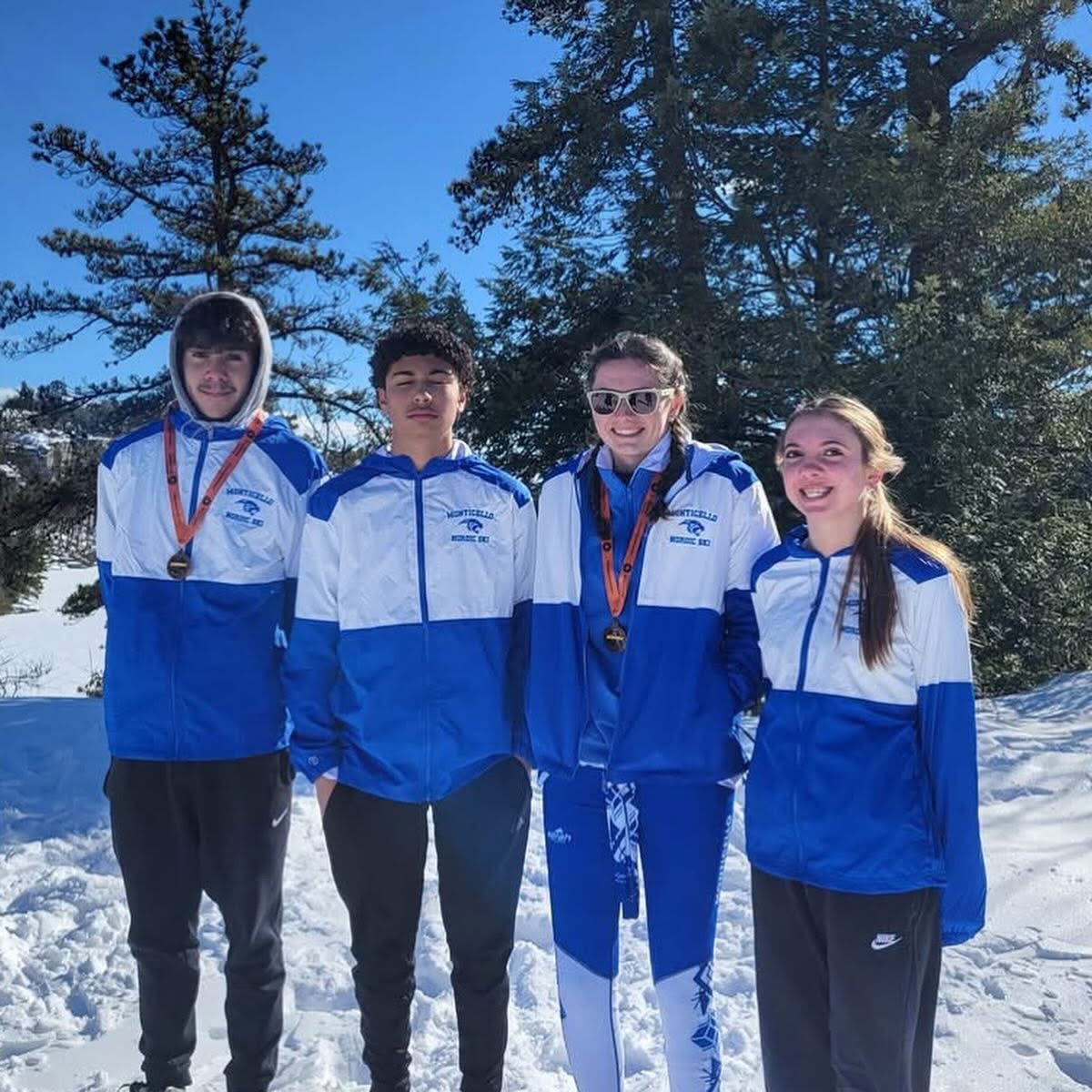 four skiiers are standing on a mountain in the snow smiling for the camera. They are dressed in Monticello windbreakers. 