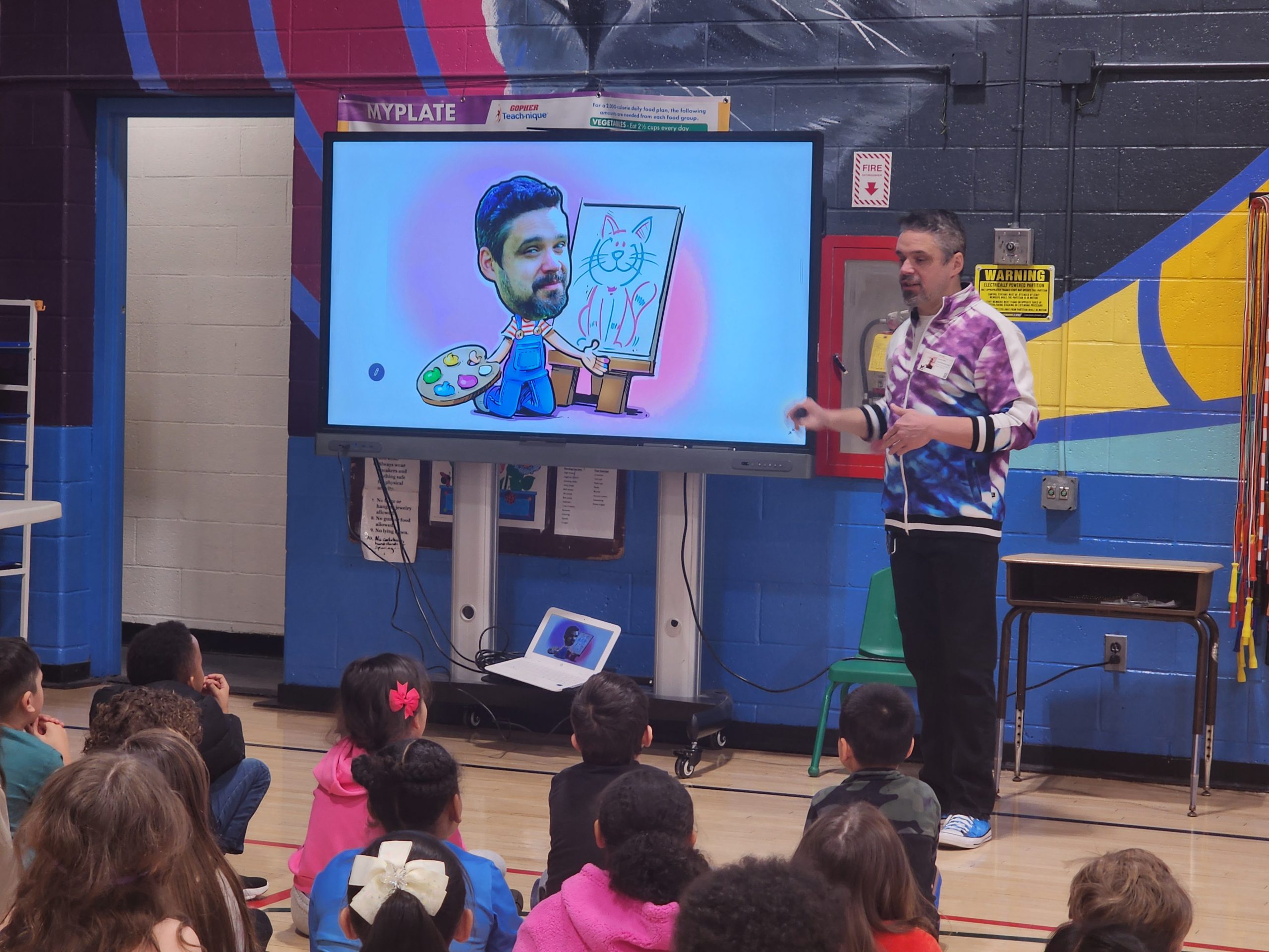 author Daniel Jude Miller is standing at the front of an auditorium speaking to students. 