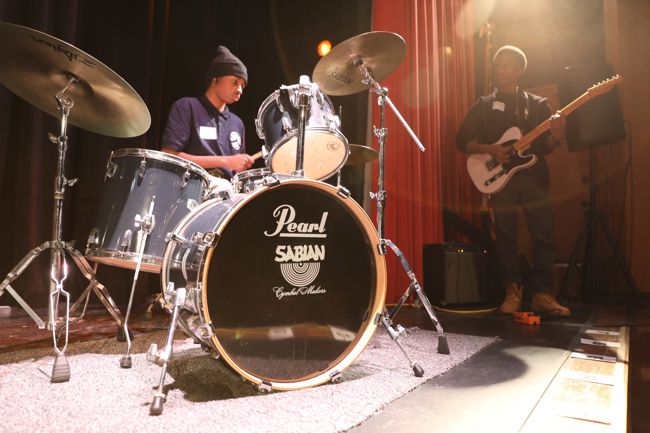 two students are playing the drums and guitar on stage.