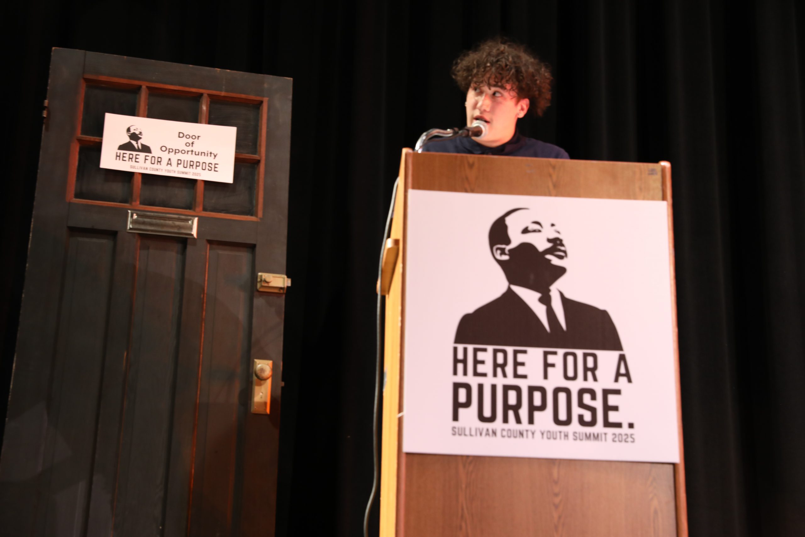 a student speaks at a podium. The podium has a sign that reads Here for a Purpose 