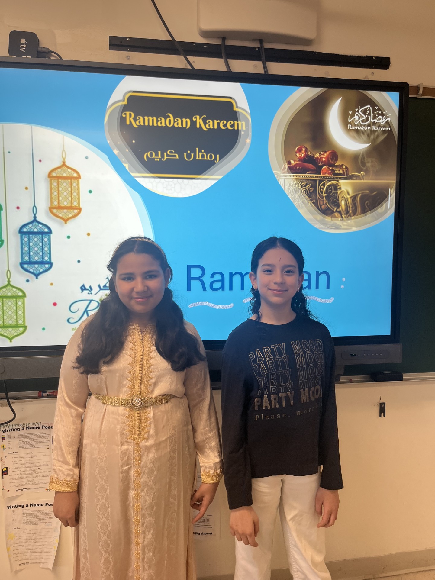 two students pose in front of a slideshow of Ramadan